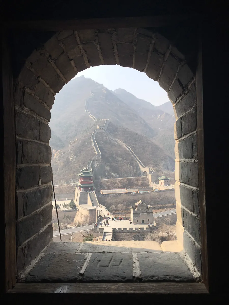 The Great Wall of China snaking up a hazy mountain ridge past a gate tower, framed through the stone-arch window of a watchtower near Beijing.