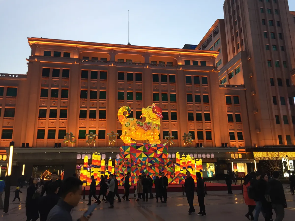 The Beijing Department Store on Wangfujing Street lit warm gold at dusk, with a big colourful zodiac lantern sculpture glowing on the plaza in front.