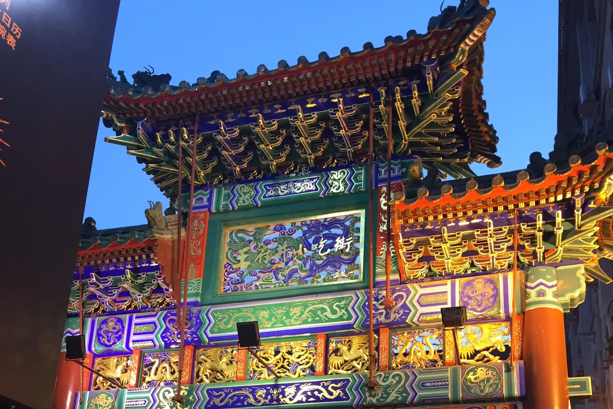 The ornately painted and gilded paifang archway of Wangfujing Snack Street in Beijing, lit up in blue, green, gold and red against a deep dusk sky.