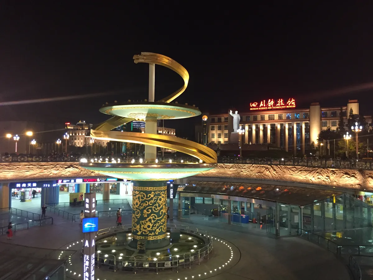 Tianfu Square in Chengdu at night: the illuminated golden spiral sculpture at the plaza's centre, with the lit Sichuan Science and Technology Museum behind and a metro entrance in the foreground.