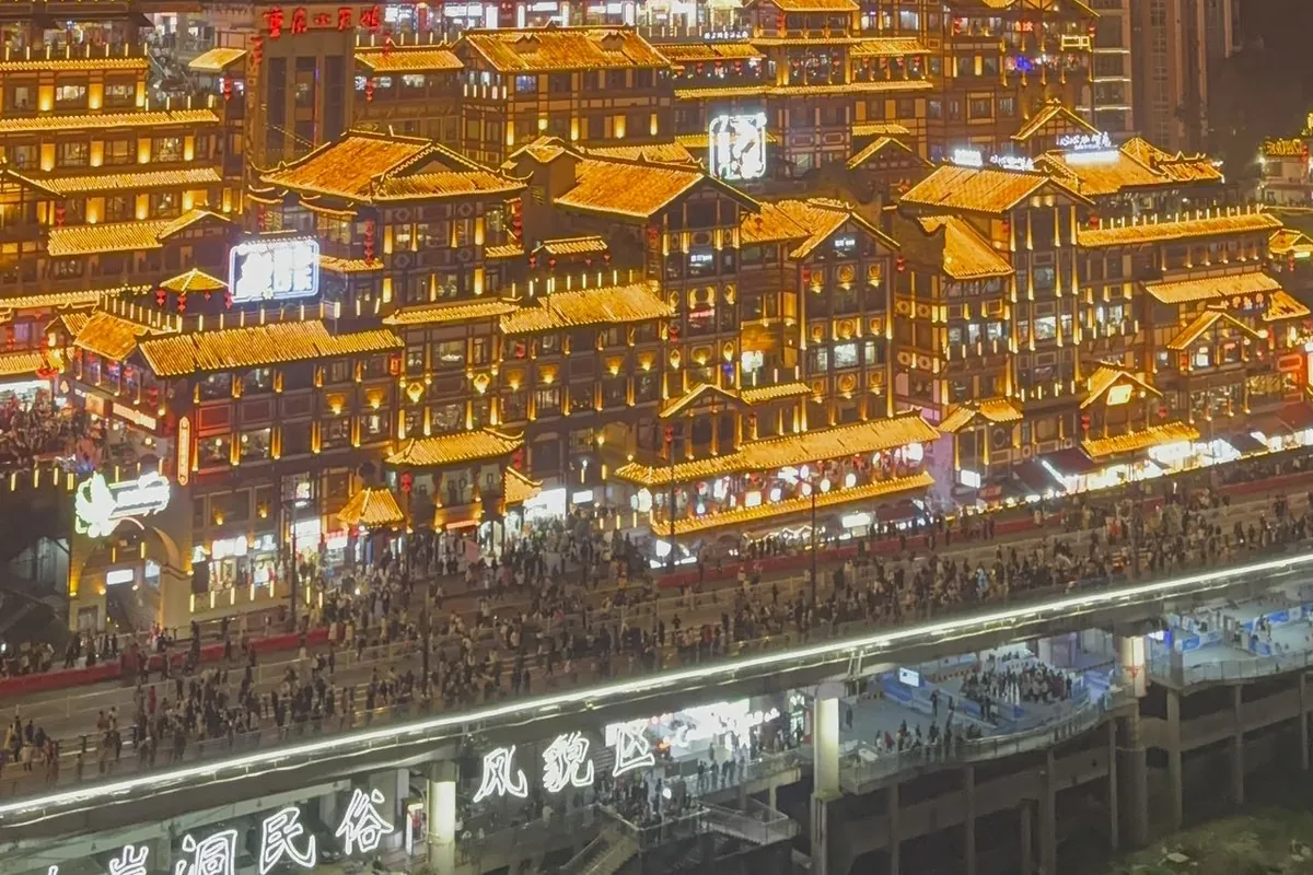 The Hongya Cave (Hongyadong) stilt-house complex in Chongqing lit gold at night, its tiered traditional rooftops stacked up the riverbank above the crowds on the waterfront below.