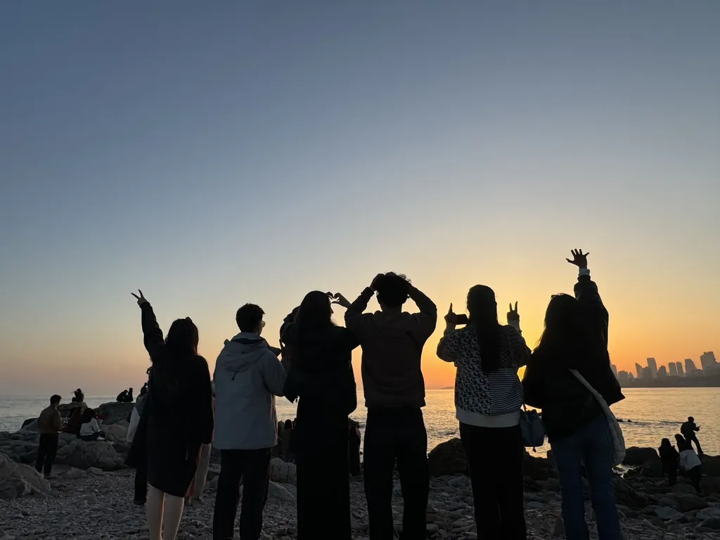 A group of friends silhouetted against a golden sunset over the sea in Dalian, making heart and peace-sign shapes with their hands.