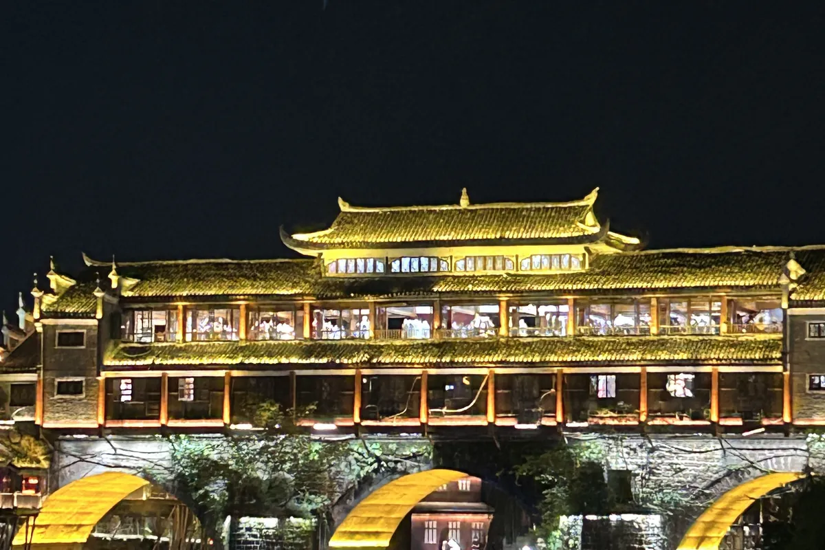 The Hong Bridge, a golden-lit covered wind-and-rain bridge, spanning the Tuo River in Fenghuang Ancient Town at night, its arches and lights mirrored in the water.