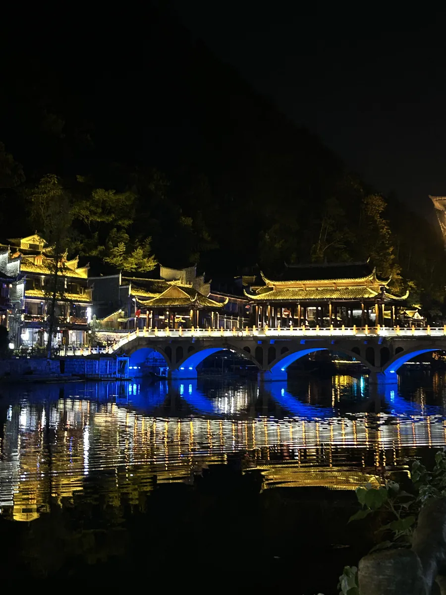 Fenghuang ancient town at night — a blue-lit arched bridge and golden stilt houses mirrored in the calm Tuo River.