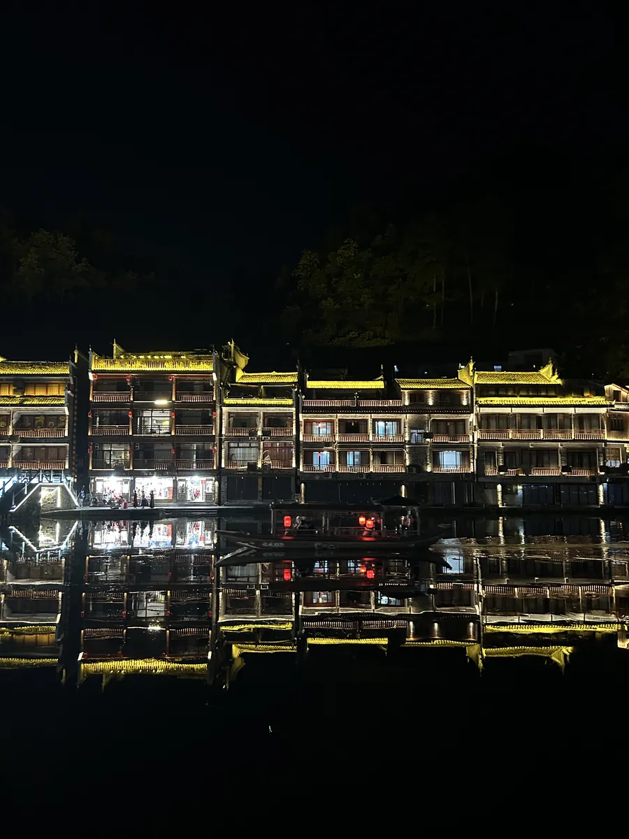 Golden-lit wooden stilt houses (diaojiaolou) leaning over the Tuo River in Fenghuang, their warm reflections shimmering on the dark water.