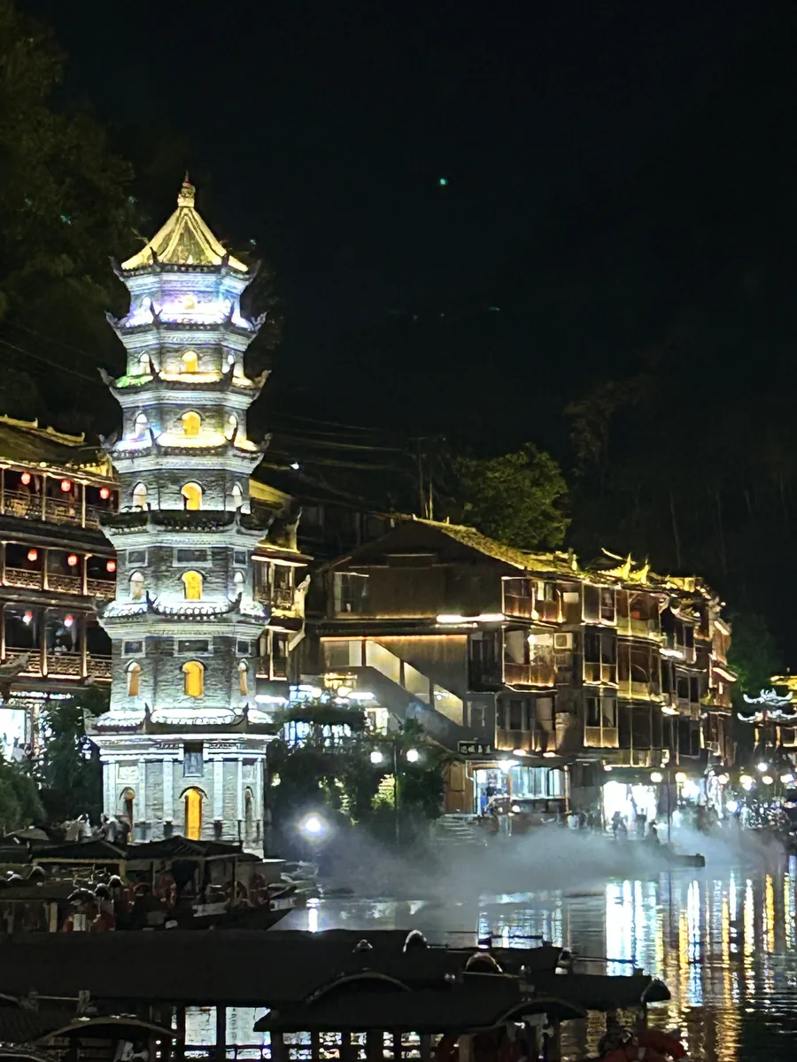 The illuminated Wanming Pagoda rising above the stilt houses and misty river in Fenghuang at night.