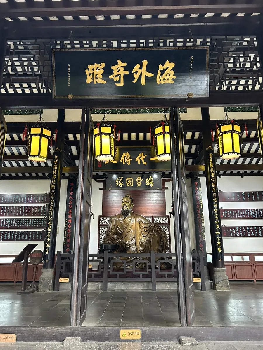 The dim shrine hall of the Dongpo Academy — a bronze statue of Su Dongpo seated beneath glowing yellow lanterns, the walls covered in calligraphy.