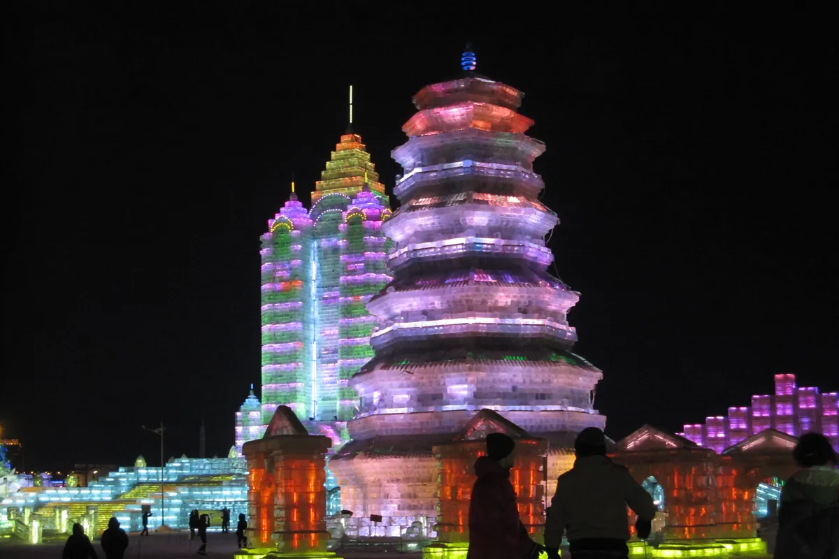 Colorful illuminated ice towers at Harbin Ice and Snow World at night, visitors silhouetted in the foreground.