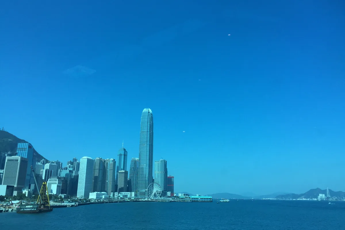 The Hong Kong Island skyline — the IFC tower and the Observation Wheel — seen across Victoria Harbour under a clear blue sky, with Victoria Peak rising behind and hills fading into the distance.