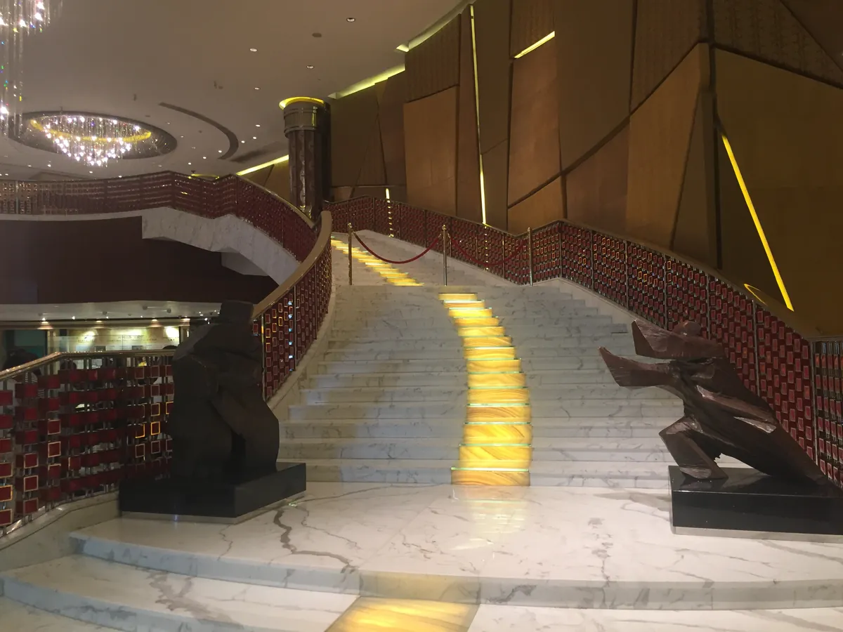 The opulent lobby of a Macau casino — a sweeping marble staircase edged in red and gold beneath a glittering chandelier.