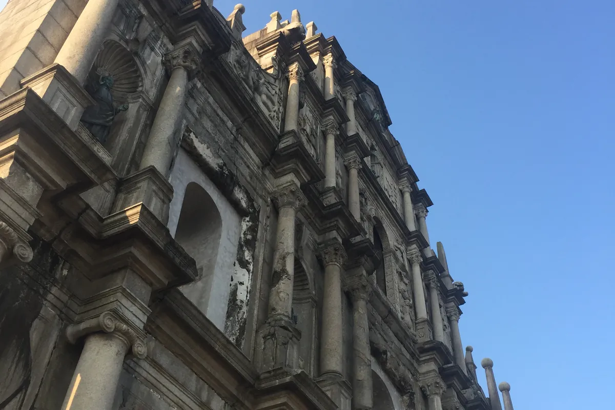 The ornate stone facade of the Ruins of St. Paul's in Macau, seen from below against a clear blue sky, its carved columns and niche statues lit by late sun.