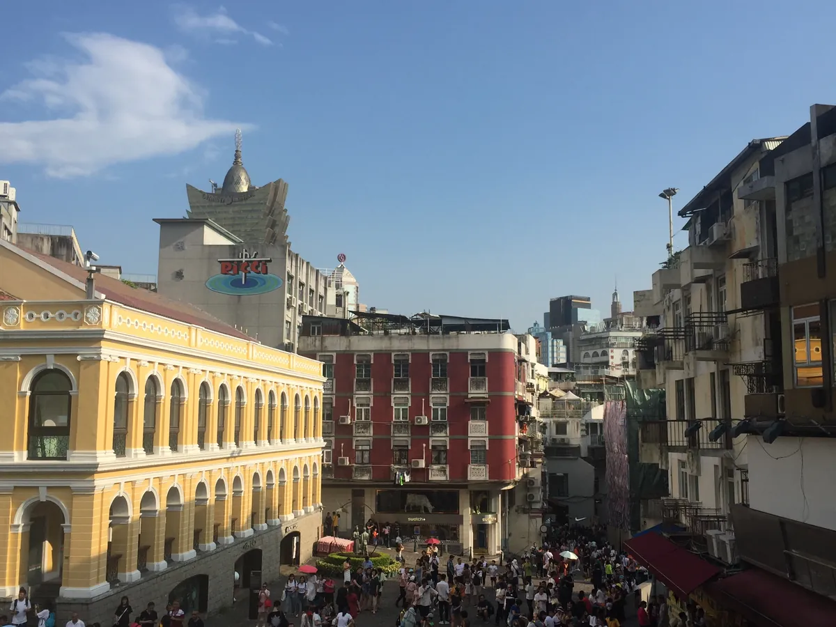 Senado Square in Macau, its pastel Portuguese colonial buildings and wave-patterned paving busy with visitors under a blue sky.