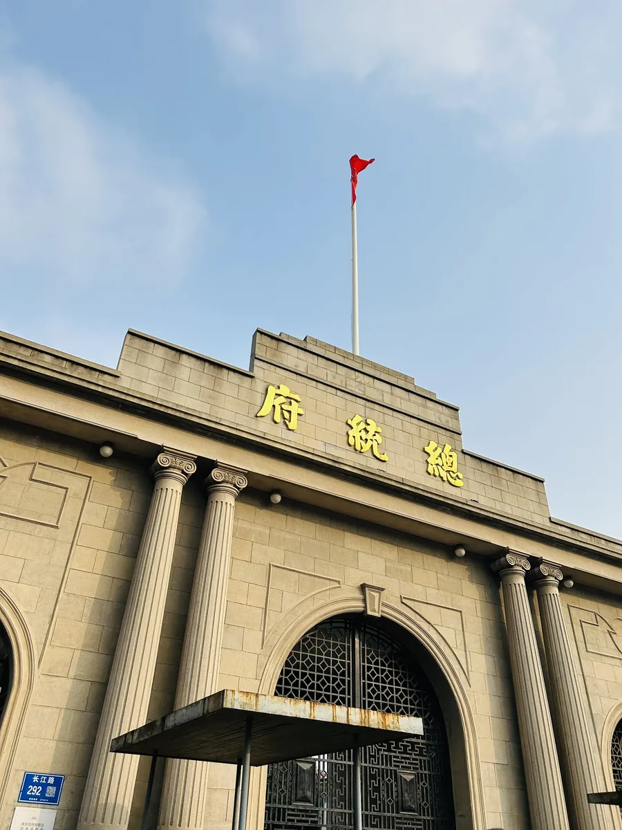 The stone facade of the Presidential Palace (Zongtongfu) in Nanjing under a blue sky, a national flag flying above its arched gateway.