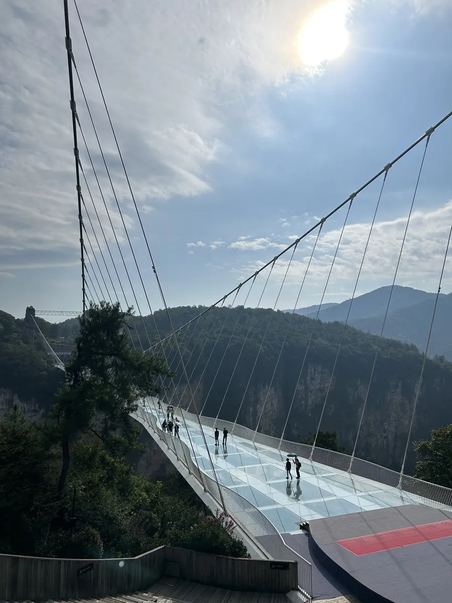 The Zhangjiajie Grand Canyon Glass Bridge — a glass-bottomed suspension span stretching across a deep forested gorge, tiny figures out on its length.