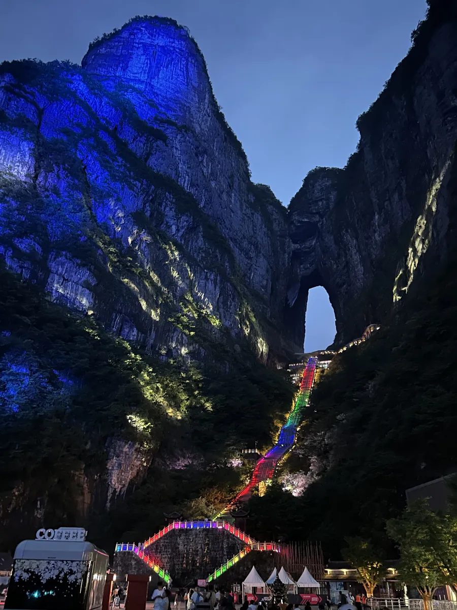 Heaven's Gate — the natural arch of Tianmen Cave — lit in rainbow colours at dusk on Tianmen Mountain in Zhangjiajie, with the illuminated staircase climbing the cliff to the arch.